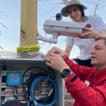 Two people checking wires in an outdoor electrical panel.