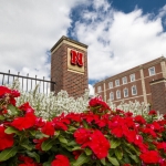 A tall brick column with the Husker "N" logo, with red and white flowers planted in front of the column.