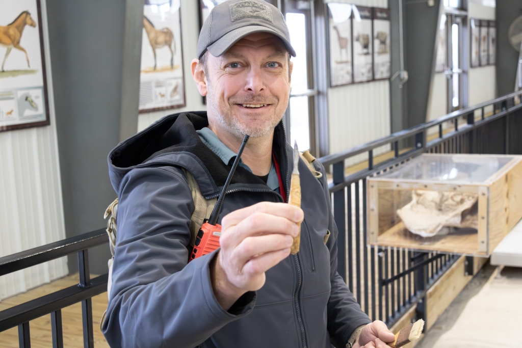 Sam Matson smiling as he holds a small, slender excavation tool toward the camera inside the Ashfall Fossil Beds visitor area.