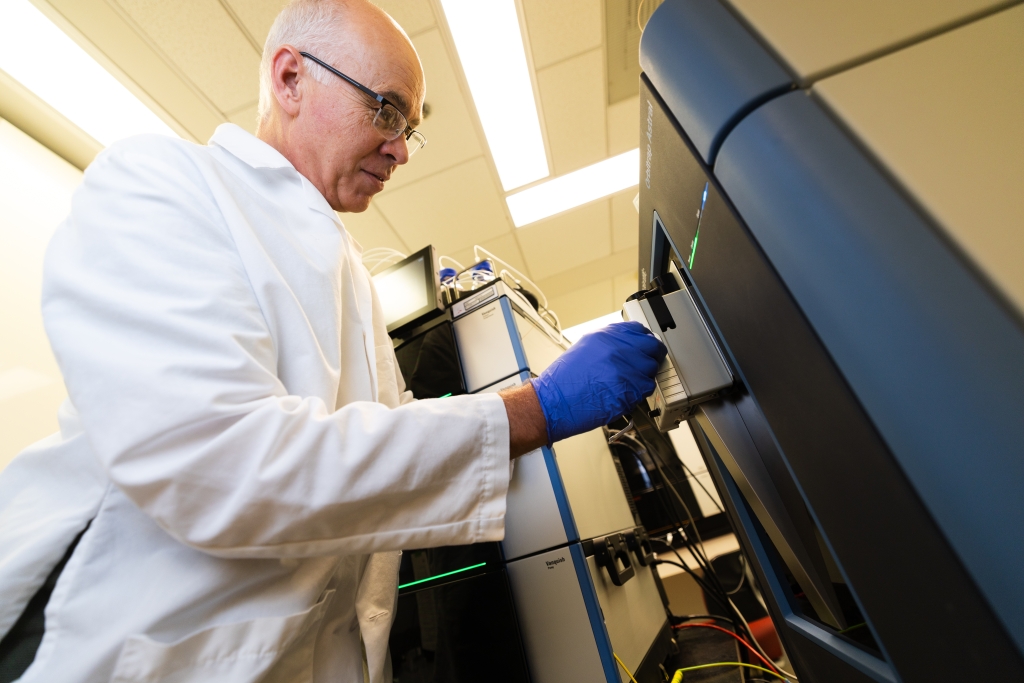 A scientists in a white lab coat and blue gloves looks at the front of a piece of equipment.