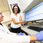 A woman with chin-length brown hair and glasses stands and uses a pen to point toward a spike in a graph on a computer screen as a seated man in glasses, a white lab coat and blue gloves uses the mouse.