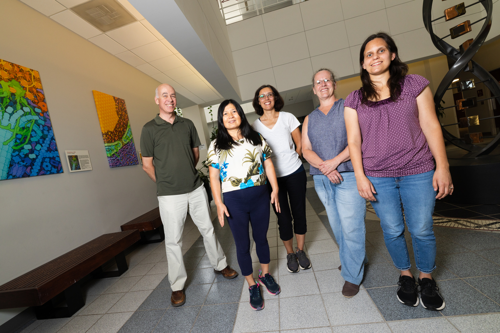 Five scientists stand stand in the lobby of a building. They are all smiling. In the background are colorful art and a statue of a strand of DNA.