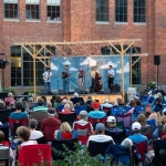 A bluegrass band performs for a large crowd of onlookers, sitting outside in camp chairs and facing the stage.