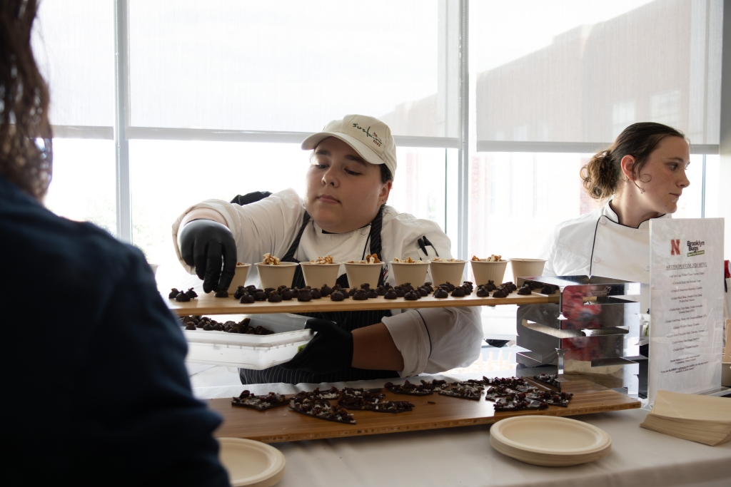 Two culinary students in chef whites plating rows of chocolate bonbons and nut-topped paper cups behind a Brooklyn Bugs menu card.