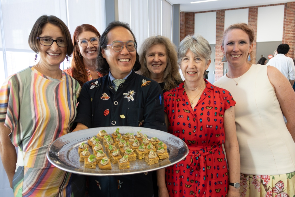 Chef Joseph Yoon holding a large platter of canapés, surrounded by five smiling women, several wearing insect-print clothing.