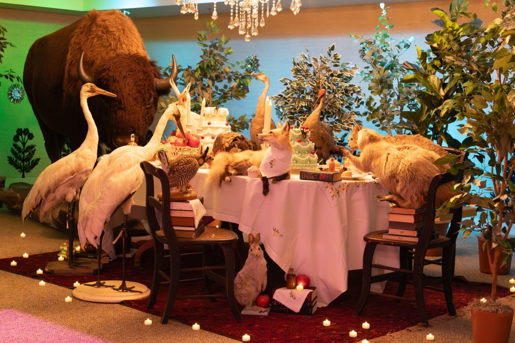 A candlelit installation of a formal dinner table set with taxidermy animals — a fox, cranes, a bison and a rabbit — gathered around a tiered cake.