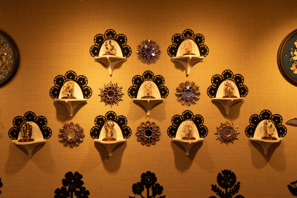 A gold gallery wall arranged like patterned wallpaper, with bell-jar shelves of insect specimens alternating with starburst medallions made of beetles.