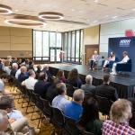An audience sits in rows of chairs facing a raised stage during a panel discussion hosted by the National Strategic Research Institute at the University of Nebraska. Three panelists sit in blue armchairs onstage in front of blue NSRI banners, with the university’s red “N” logo visible on the wall behind them. The modern conference room features large windows, wood flooring, and circular ceiling lights.