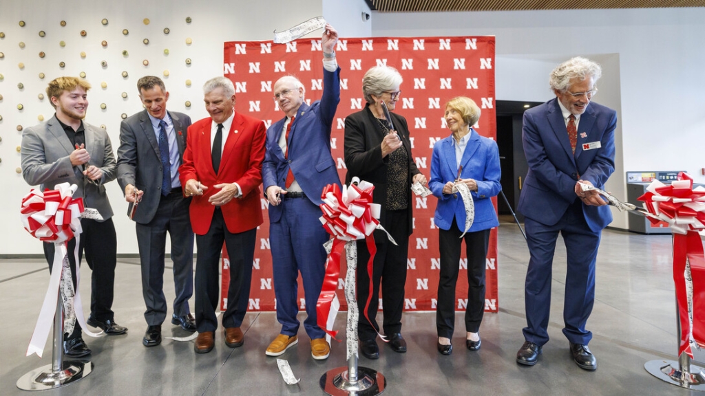 A group of seven people stands in a bright indoor space during a ribbon-cutting ceremony. Several participants hold oversized scissors and sections of a white ribbon with large red bows, while smiling and celebrating in front of a backdrop patterned with red University of Nebraska “N” logos. Attendees wear business attire, including blue, black, and red jackets, and one person raises a piece of ribbon overhead. The event appears to mark the opening or dedication of a new space.