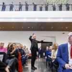 A large group of people gathers in a modern indoor space, with several seated in rows and others standing along an upper-level balcony. At the center, a man in dark clothing stands and gestures toward the ceiling while speaking to the group. In the foreground, attendees listen, including a woman in black on the left and a man in a blue suit with a red tie on the right. The room features wood slat ceilings, bright white walls, and colorful artwork.