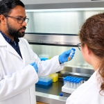 A scientist wearing a white lab coat, safety glasses, and blue gloves demonstrates a small sample tube to a colleague in a white lab coat inside a laboratory. The scientist holds a tube containing an orange-colored liquid, while racks of blue-capped sample tubes and laboratory supplies sit on the work surface behind them. They stand in front of a laboratory fume hood with a sign reading “KEEP THIS CLOSED ALWAYS when not in use.”