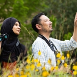 A woman in a black hijab and blazer looks at a man to her right as she holds up a pair of binoculars. The man wears a blue and yellow plaid shirt and has his left hand in the air with his pointer finger up. He holds binoculars in his right hand. They stand in a green space with tall grasses and a stand of black-eyed susans.