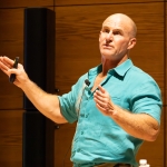 A man in a turquoise embroidered shirt speaks during a lecture, holding a presentation remote and gesturing with his hands. He stands in front of a warm wood-paneled wall, with a black speaker behind him and a microphone on a stand to his right.